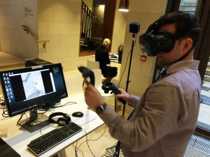 Man with VR headset stands in front of computer monitor with VR controllers in hand.